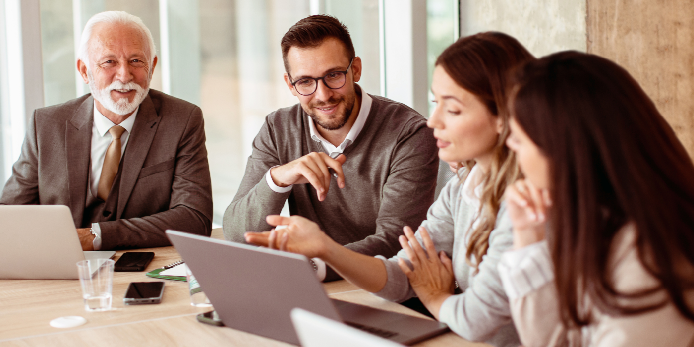 Quatro pessoas sentadas em uma mesa de trabalho de frente para dois computadores portáteis, laptops. Dois homens, um de cabelo e barba brancos e outro de cabelo preto e usando óculos, e duas mulheres, ambas de cabelo preto.